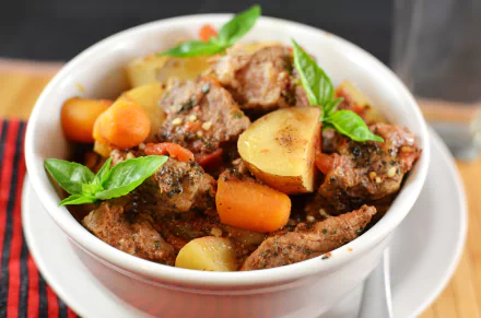 Close-up 4K Ultra HD image of a hearty stew with tender meat, carrots, potatoes, and fresh basil leaves in a white bowl on a wooden table.