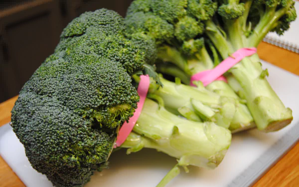 Close-up of three fresh broccoli heads bound with pink rubber bands on a cutting board — vibrant 4K Ultra HD food image, PC desktop wallpaper and background.