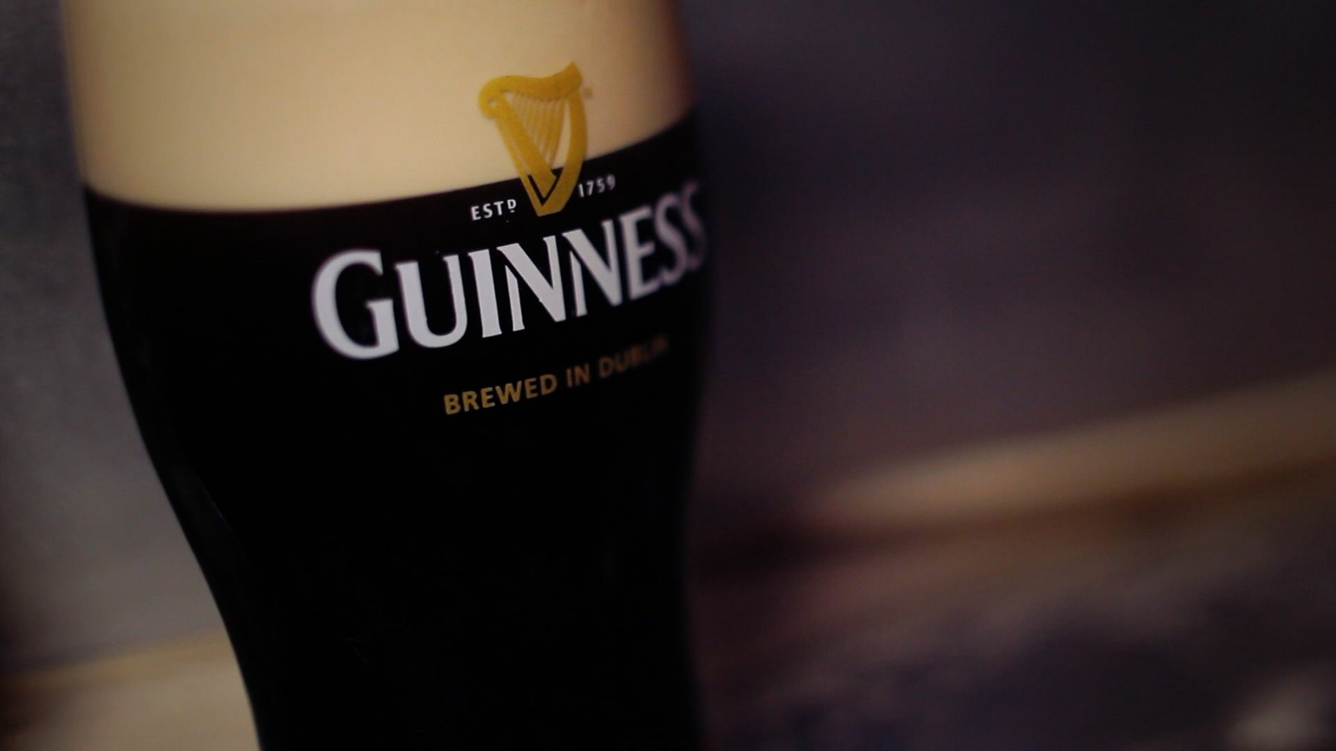 Close-up HD wallpaper of a man-made Guinness pint glass filled with dark beer and creamy foam on top against a blurred background.