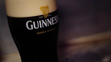 Close-up HD wallpaper of a man-made Guinness pint glass filled with dark beer and creamy foam on top against a blurred background.