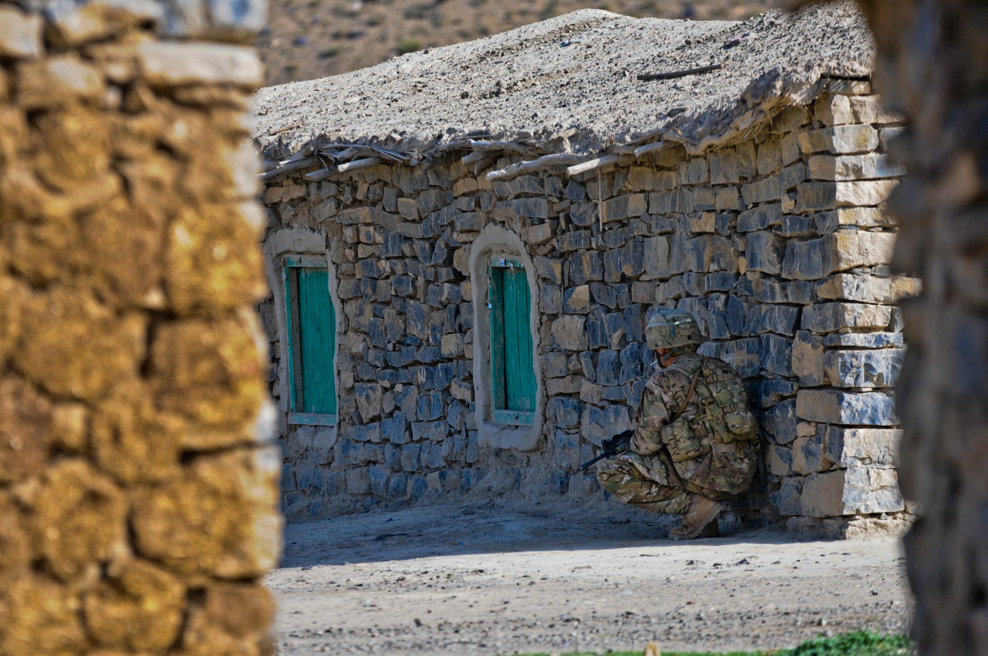 4K Ultra HD image of a camouflaged soldier crouching beside a stone building with green windows in a military setting.