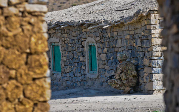 4K Ultra HD image of a camouflaged soldier crouching beside a stone building with green windows in a military setting.