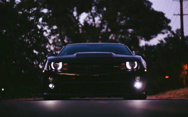 Front view of a black Chevrolet Camaro SS muscle car with illuminated headlights, set against a dark, tree-lined backdrop in 4K Ultra HD quality.