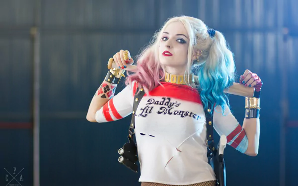 A woman with twintails, blue eyes, and lipstick cosplays as Harley Quinn, wearing a “Daddy’s Lil Monster” shirt. This HD desktop wallpaper captures her confident and colorful portrayal.