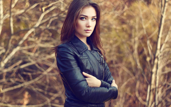 HD PC desktop wallpaper/background: woman model with long brown hair in a black leather jacket standing before tangled autumn branches, arms crossed.