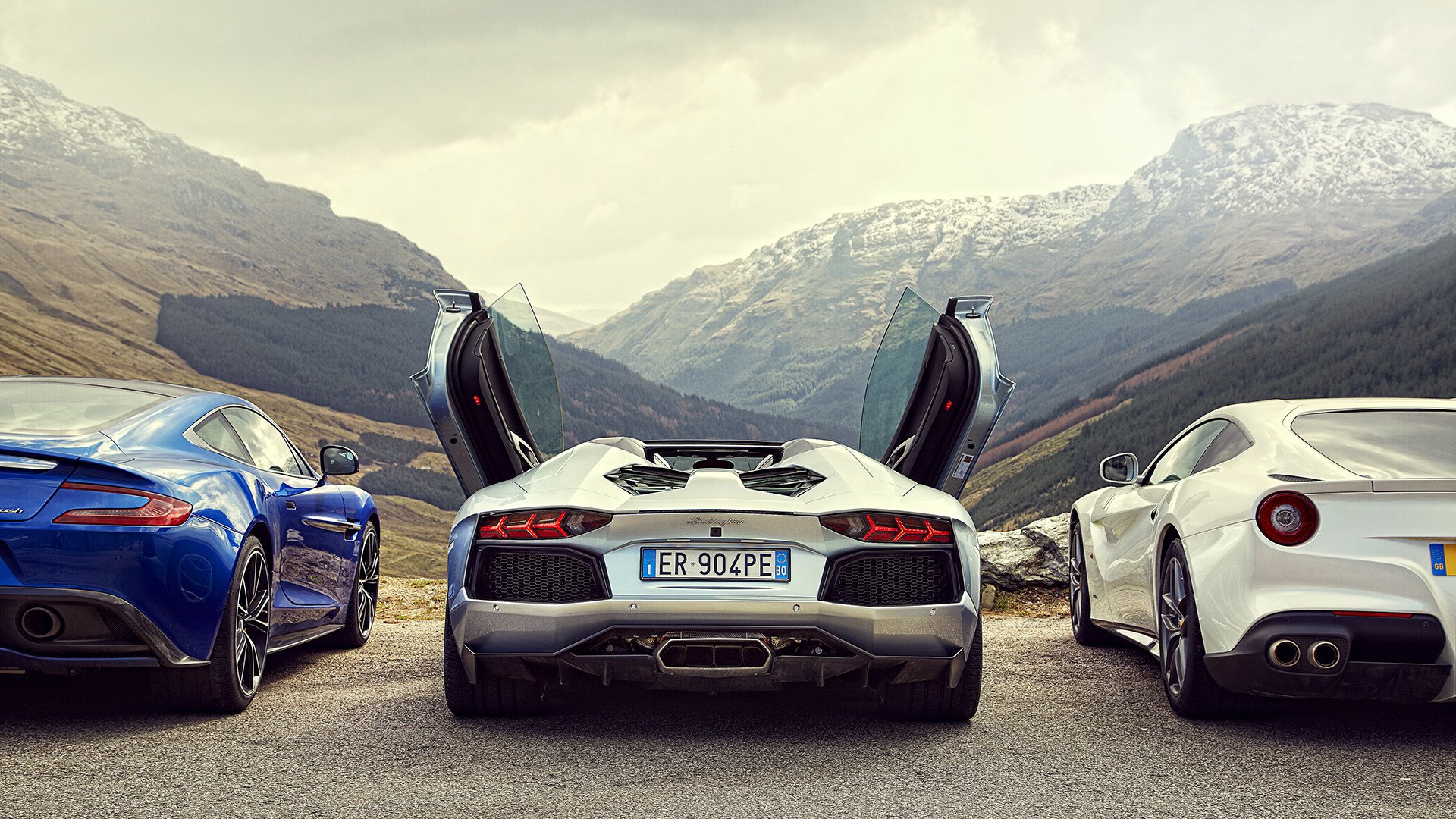 HD PC desktop wallpaper of a Lamborghini Aventador with its doors open, flanked by two other sports cars against a backdrop of mountainous terrain.