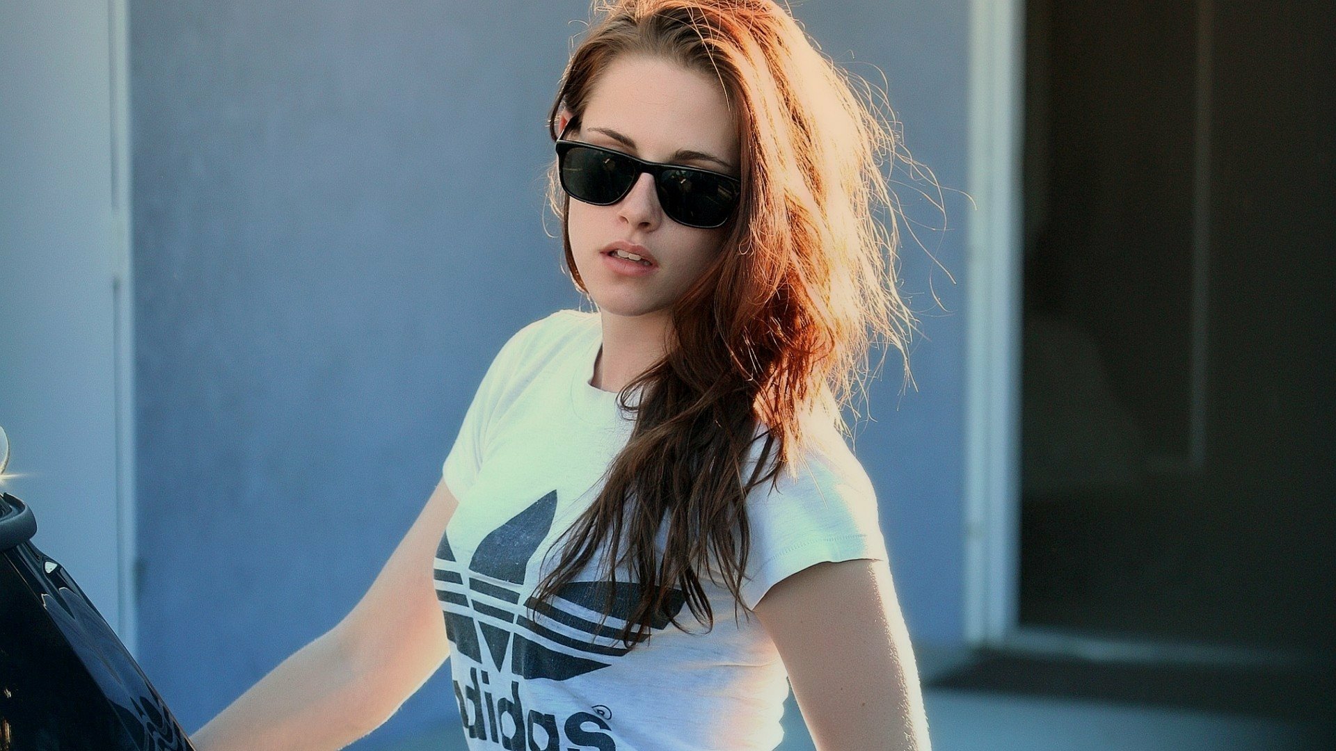 A stylish Kristen Stewart wearing sunglasses and a casual Adidas shirt stands confidently, captured in a vibrant HD wallpaper for PC desktops.