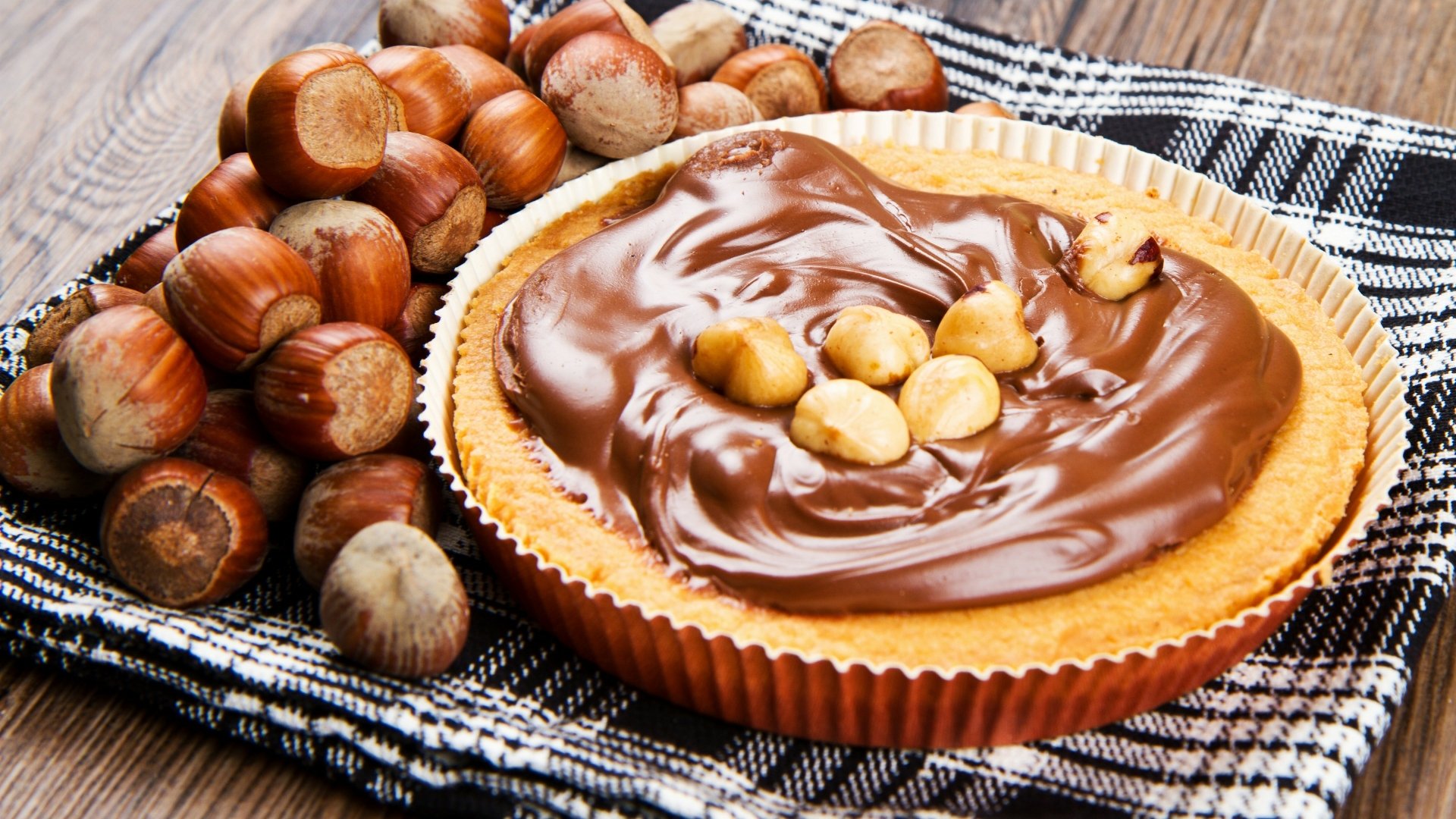 A rich chocolate hazelnut pie topped with whole hazelnuts, surrounded by fresh hazelnuts on a cloth, captured in 4K Ultra HD as a vibrant dessert desktop wallpaper.