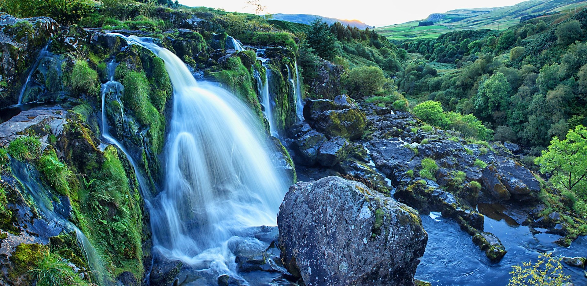 Lush green moss surrounds the cascading Loup of Fintry Waterfall flowing into a river, set in the scenic Scottish landscape captured in 4K Ultra HD detail.