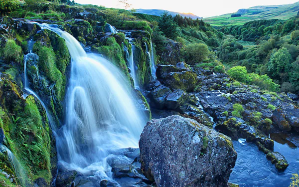 Lush green moss surrounds the cascading Loup of Fintry Waterfall flowing into a river, set in the scenic Scottish landscape captured in 4K Ultra HD detail.