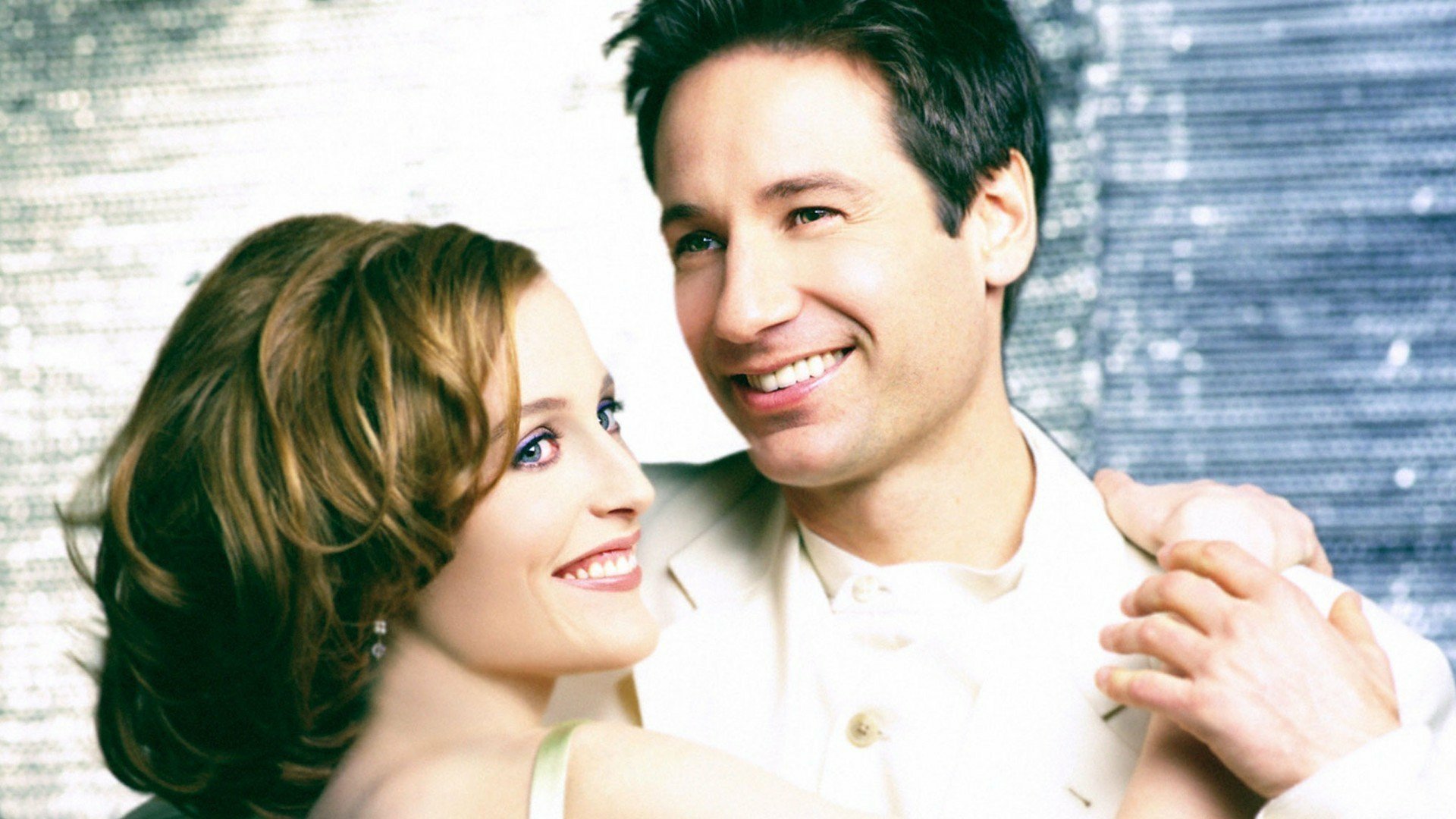 HD desktop wallpaper featuring agents Mulder and Scully from the TV show The X-Files, smiling against a blurred metallic background.
