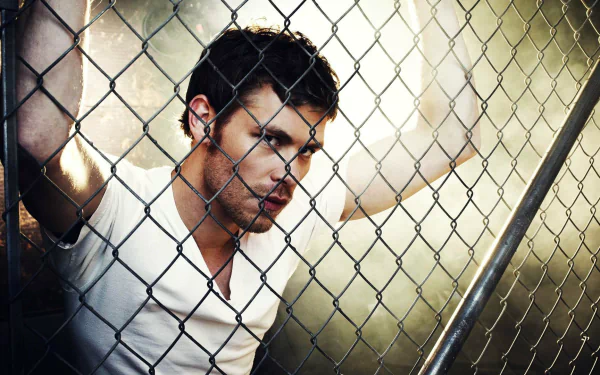 A dramatic HD wallpaper featuring a character from The Vampire Diaries, showcasing a brooding male figure behind a chain-link fence, illuminated by a moody background.