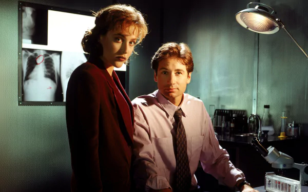 HD PC desktop wallpaper from the TV show The X-Files showing two FBI agents in a dim lab—one seated at a desk lamp, the other standing beside an X-ray on the wall.