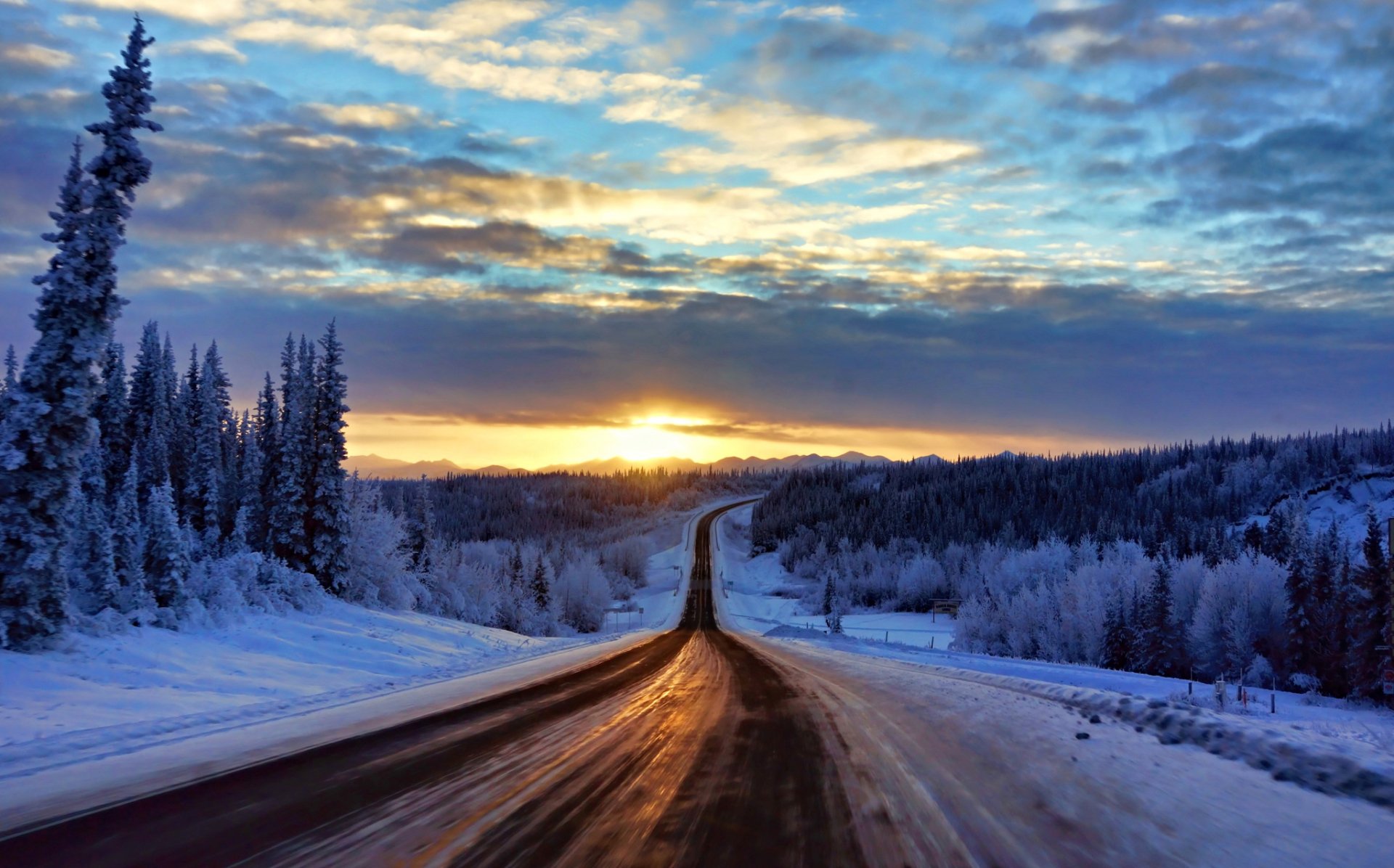 A winter landscape featuring a snow-covered forest and a man-made road stretching toward a sunrise beneath a cloud-filled sky.