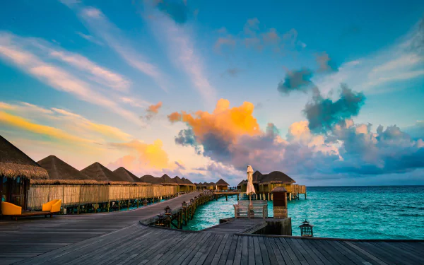 A vibrant sunset over the turquoise ocean, highlighting a wooden walkway leading to luxury bungalows in a Maldives resort, surrounded by fluffy clouds. 4K Ultra HD wallpaper.