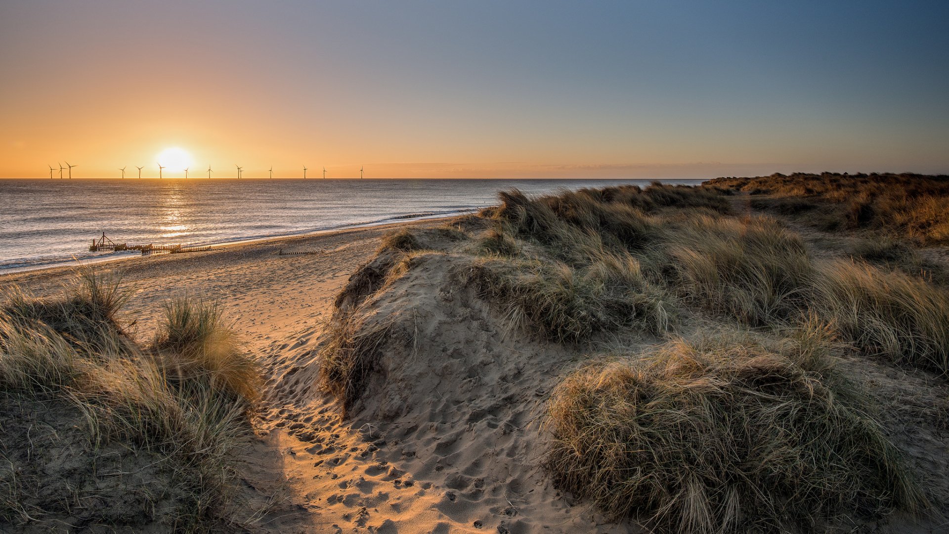 Sunrise over a sandy beach and dunes with an ocean horizon and wind turbines in the distance, captured in a serene nature landscape for HD desktop wallpaper.