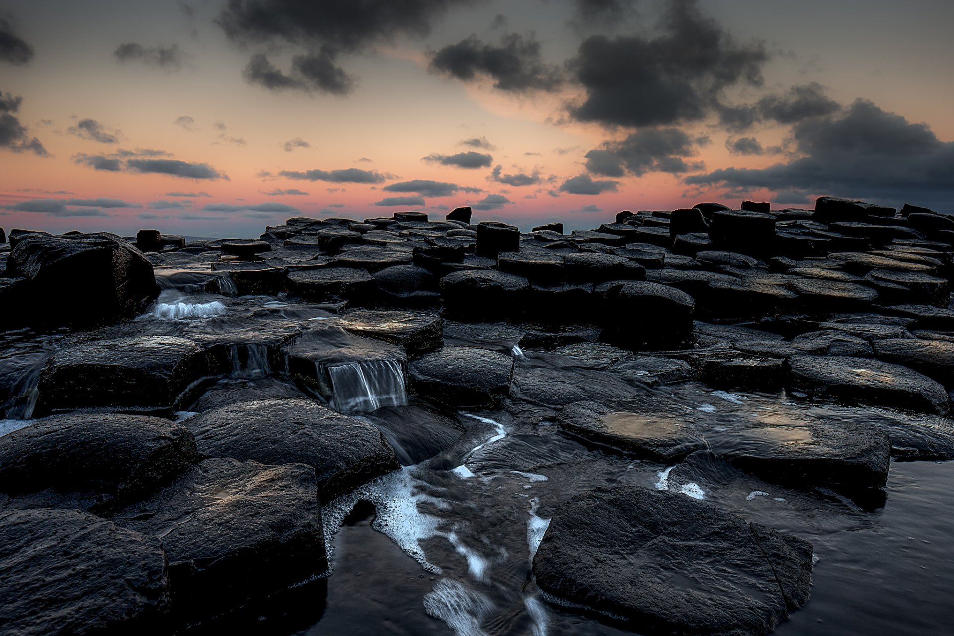 HD PC desktop wallpaper featuring a seascape with dark rocks, flowing water, and a cloudy sky during sunset, highlighting nature's serene beauty.