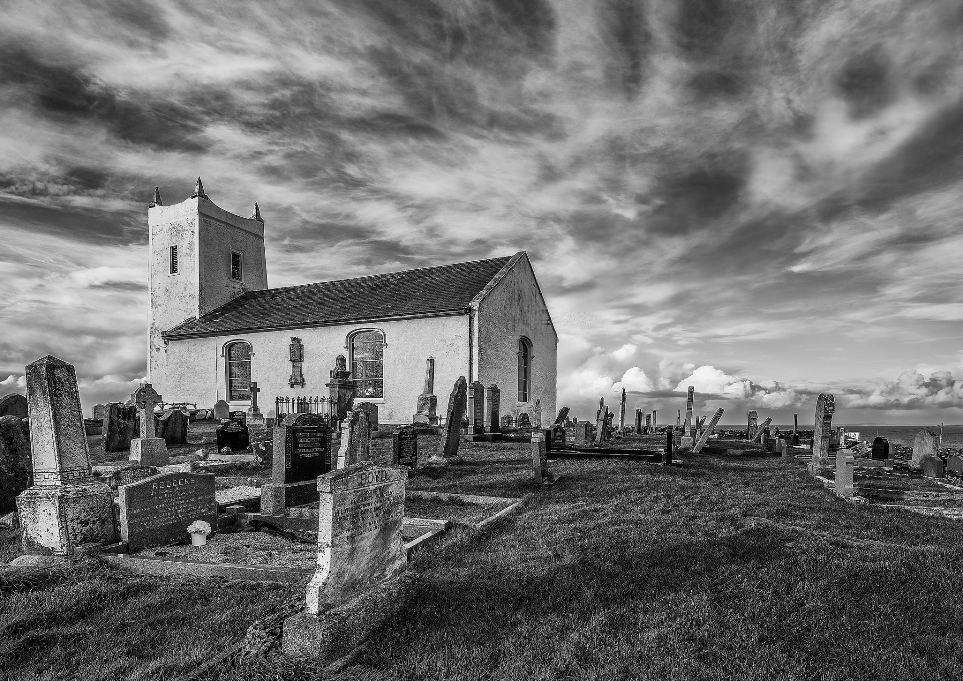 Black and white HD desktop wallpaper of a cemetery with gravestones in front of a church under a dramatic cloudy sky.