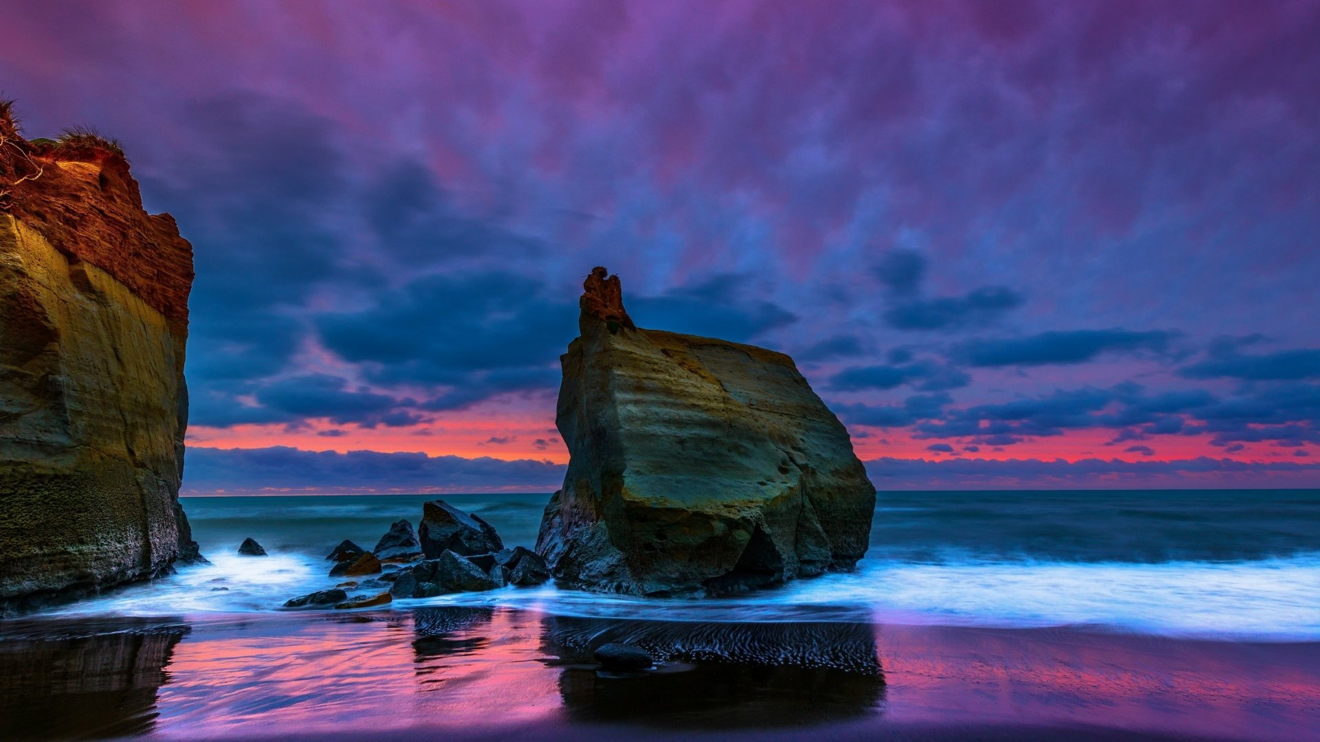 A stunning sunset over the ocean, casting vibrant colors across the sky. Rocky formations rise above gentle waves, creating a serene nature scene in 4K Ultra HD.