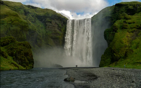 5K Ultra HD PC Desktop Wallpaper and Background: Skógafoss waterfall framed by mossy green cliffs, mist rising as water plunges into a rocky riverbed under a cloudy sky (nature).