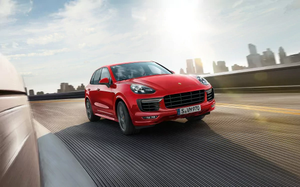 HD desktop wallpaper featuring a red Porsche Cayenne SUV driving on a highway with a city skyline in the background.