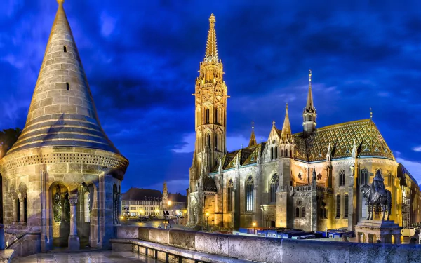 Illuminated Matthias Church with its colorful roof tiles and nearby turret tower in Budapest, Hungary, set against a deep blue evening sky.