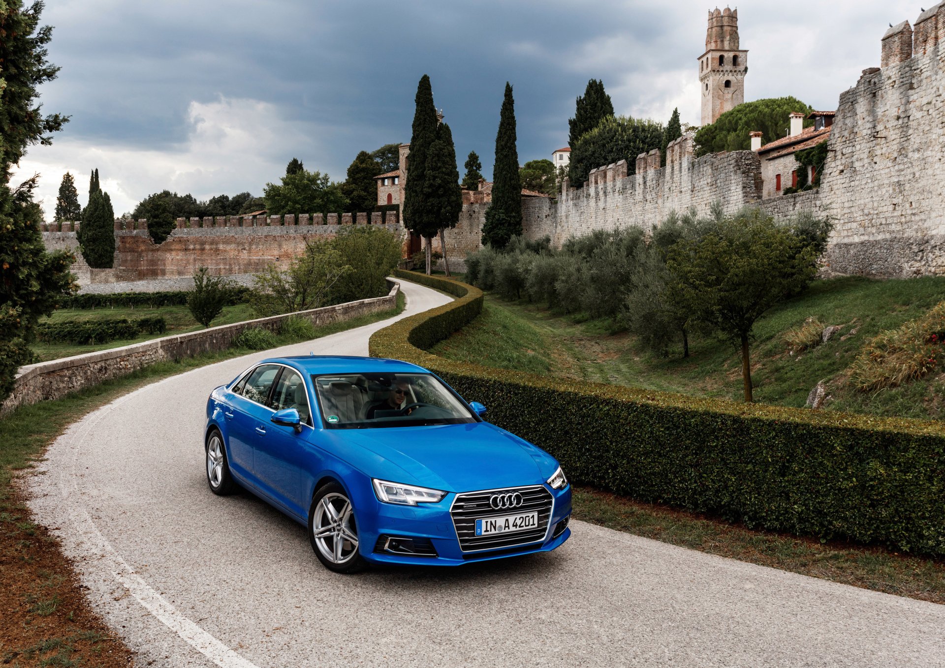 A sleek blue Audi A4 parked on a winding road, surrounded by lush greenery and historic stone walls, set against a dramatic sky. Ideal HD wallpaper for car enthusiasts.