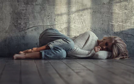 A blonde model lies against a textured wall, curled up on the floor in a cozy sweater and jeans, capturing a serene and introspective mood in a high-definition wallpaper.