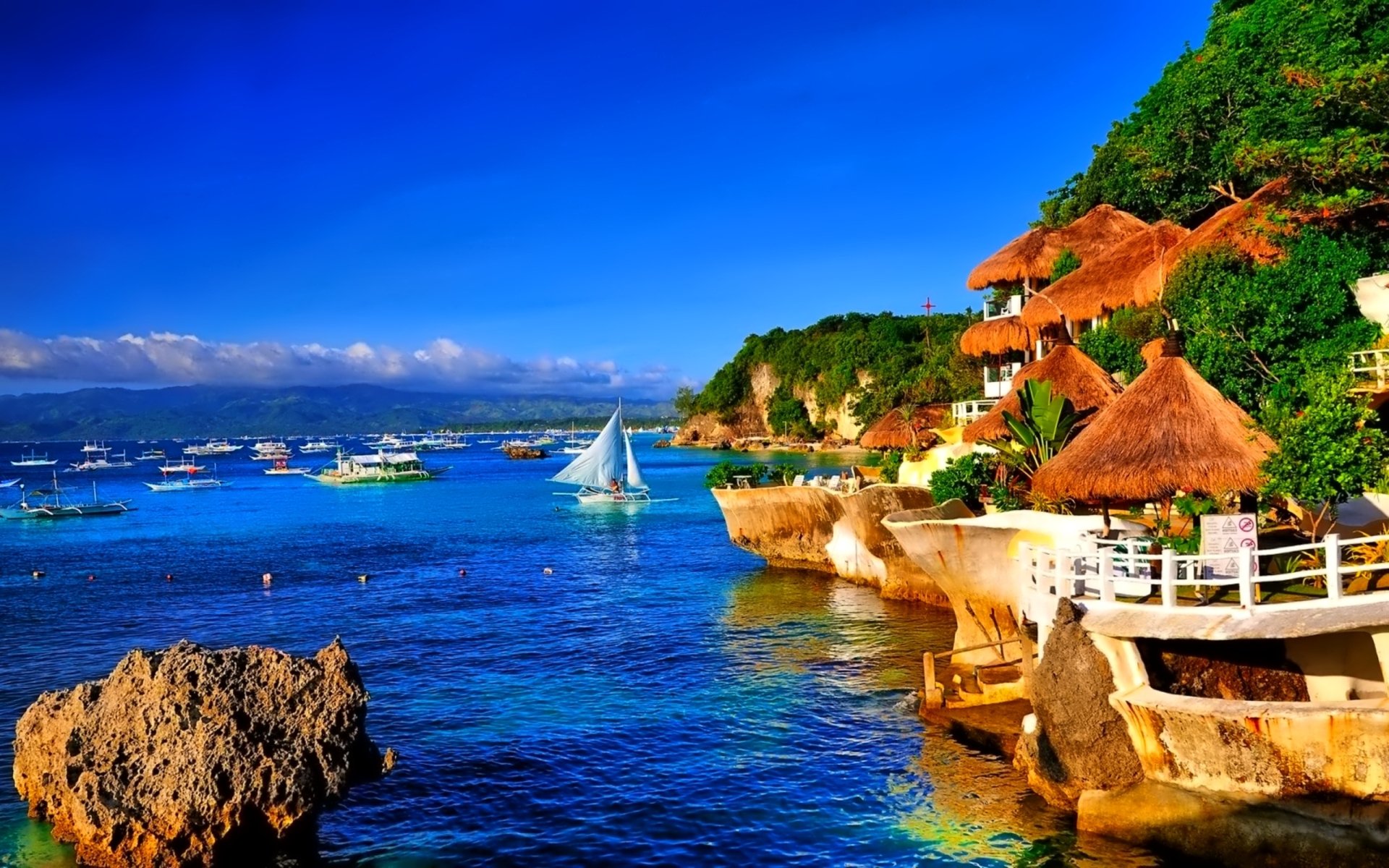 HD PC desktop wallpaper featuring a tropical coastline with vibrant ocean waters, boats, a sailboat, and thatched-roof huts and villas nestled on rocky cliffs.