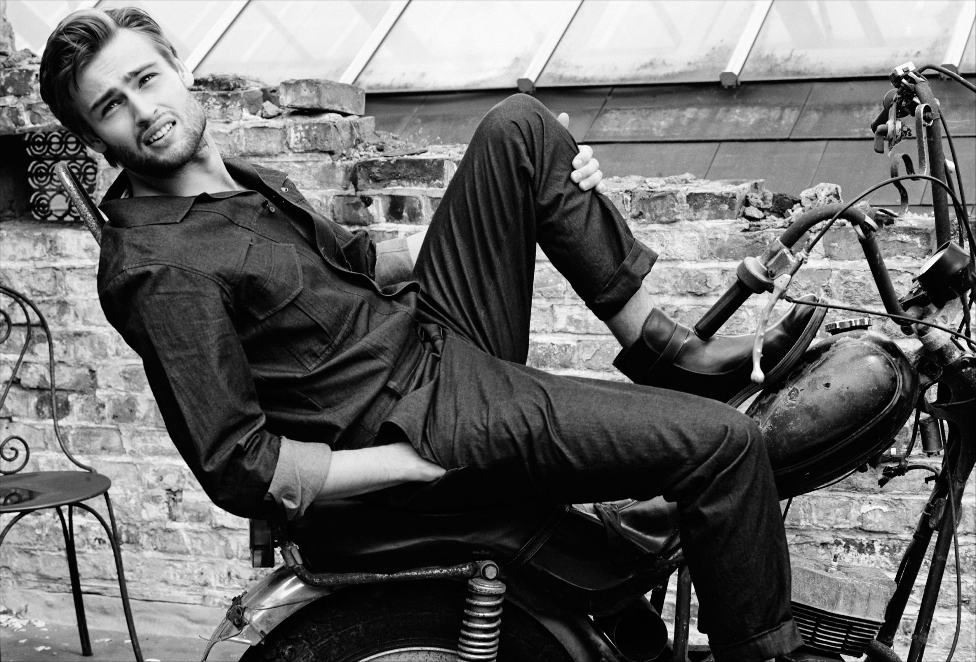Black and white HD desktop wallpaper featuring English actor Douglas Booth reclining on a vintage motorcycle against a brick wall backdrop.