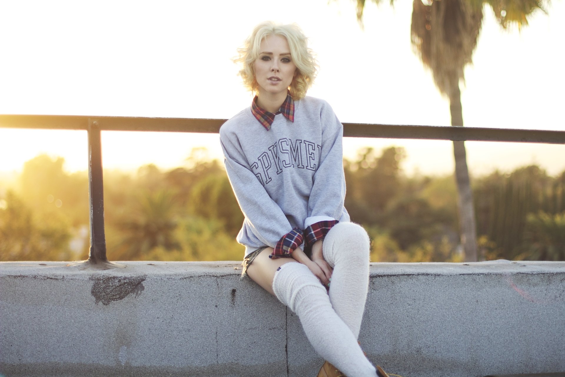 Blonde model seated on a low concrete wall at sunset, wearing an oversized sweater and thigh-high socks — 5K Ultra HD PC desktop wallpaper.