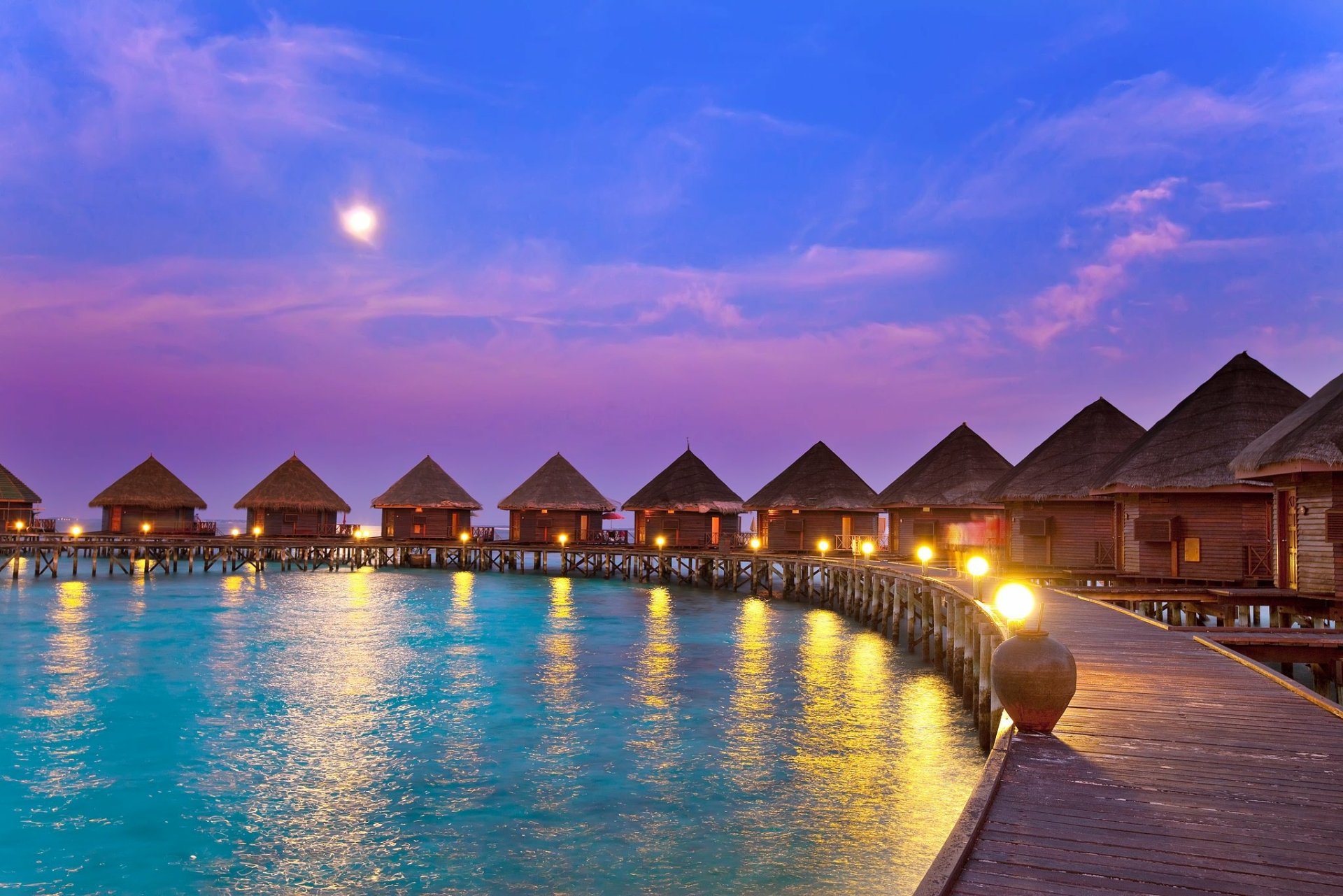 Tropical resort at sunset with overwater bungalows along a curved wooden walkway, glowing lights reflecting on calm water under a vibrant evening sky.