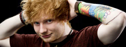 Ed Sheeran in a maroon shirt, showcasing his colorful arm tattoo. He is posing with his hands behind his head, against a black background. HD desktop wallpaper and background.
