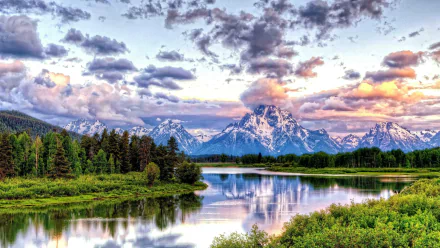 HD desktop wallpaper shows a serene river reflecting lush greenery and snow-capped mountains under a dramatic sky filled with clouds in Grand Teton National Park. Beautiful nature landscape.