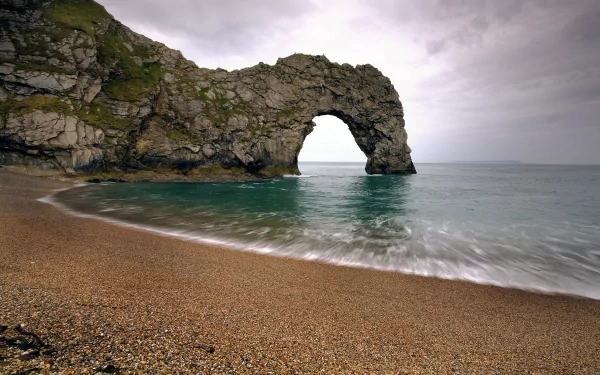 HD desktop wallpaper featuring Durdle Door, a natural limestone arch on a rocky cliff by the beach, with calm sea waves under a cloudy sky.