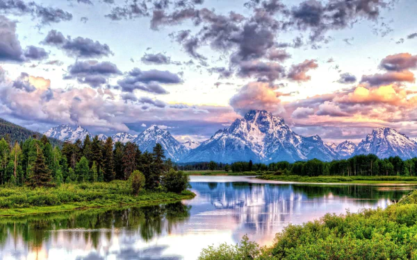 HD desktop wallpaper shows a serene river reflecting lush greenery and snow-capped mountains under a dramatic sky filled with clouds in Grand Teton National Park. Beautiful nature landscape.