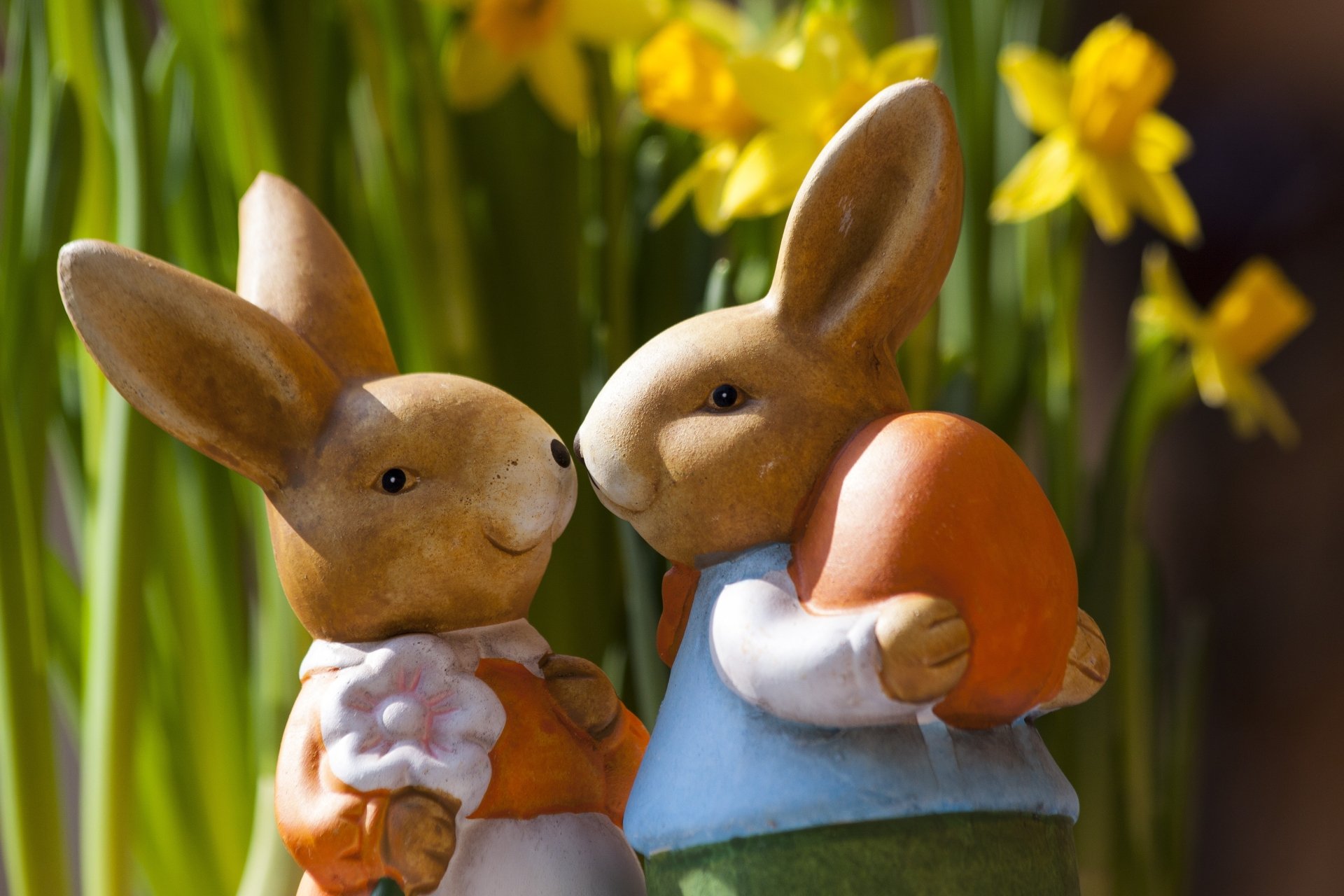 Two ceramic rabbits holding Easter eggs with yellow daffodils in the background, captured in vibrant 4K Ultra HD for a festive holiday desktop wallpaper.
