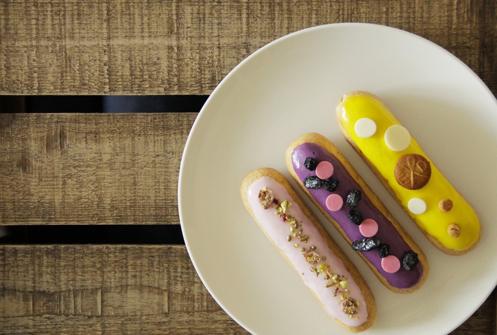 4K Ultra HD PC desktop wallpaper showing three colorful éclair pastries (pink, purple, yellow), a dessert topped with berries, nuts and chocolate on a white plate over a wooden table.