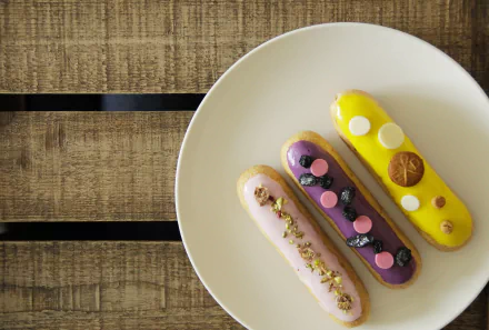 4K Ultra HD PC desktop wallpaper showing three colorful éclair pastries (pink, purple, yellow), a dessert topped with berries, nuts and chocolate on a white plate over a wooden table.