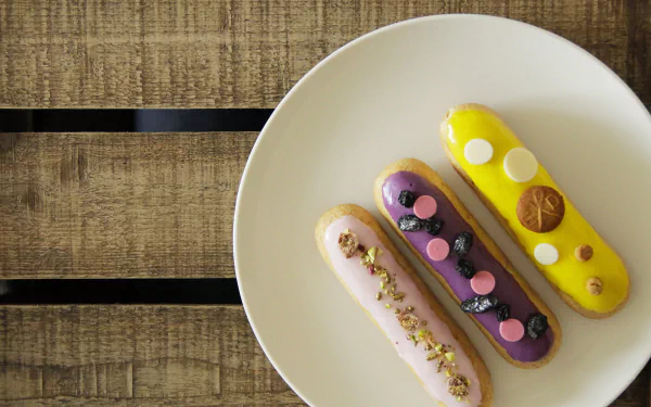 4K Ultra HD PC desktop wallpaper showing three colorful éclair pastries (pink, purple, yellow), a dessert topped with berries, nuts and chocolate on a white plate over a wooden table.