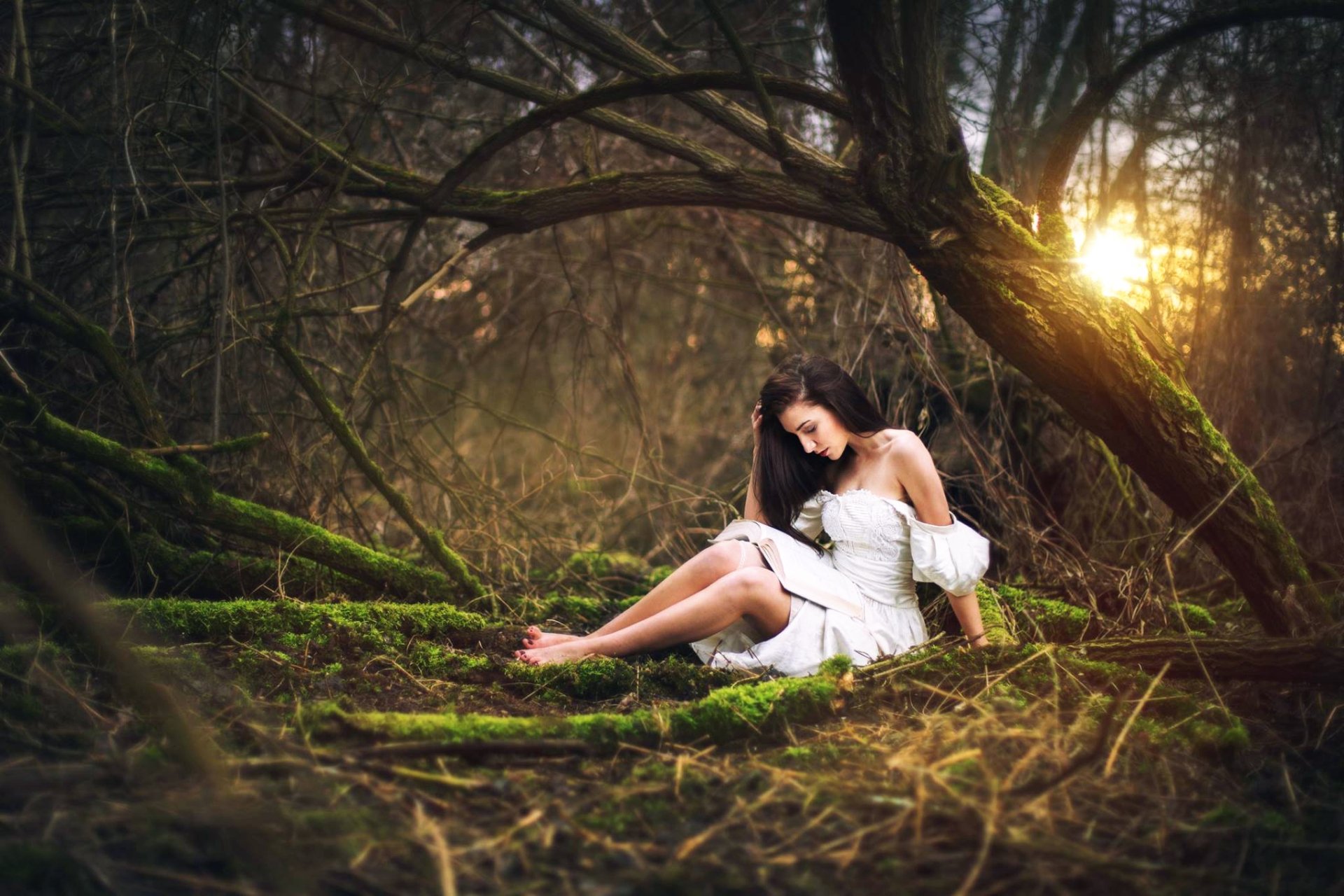 A brunette woman in a white dress sits barefoot on mossy ground in a forest, bathed in warm sunlight, creating a serene and moody HD desktop wallpaper.