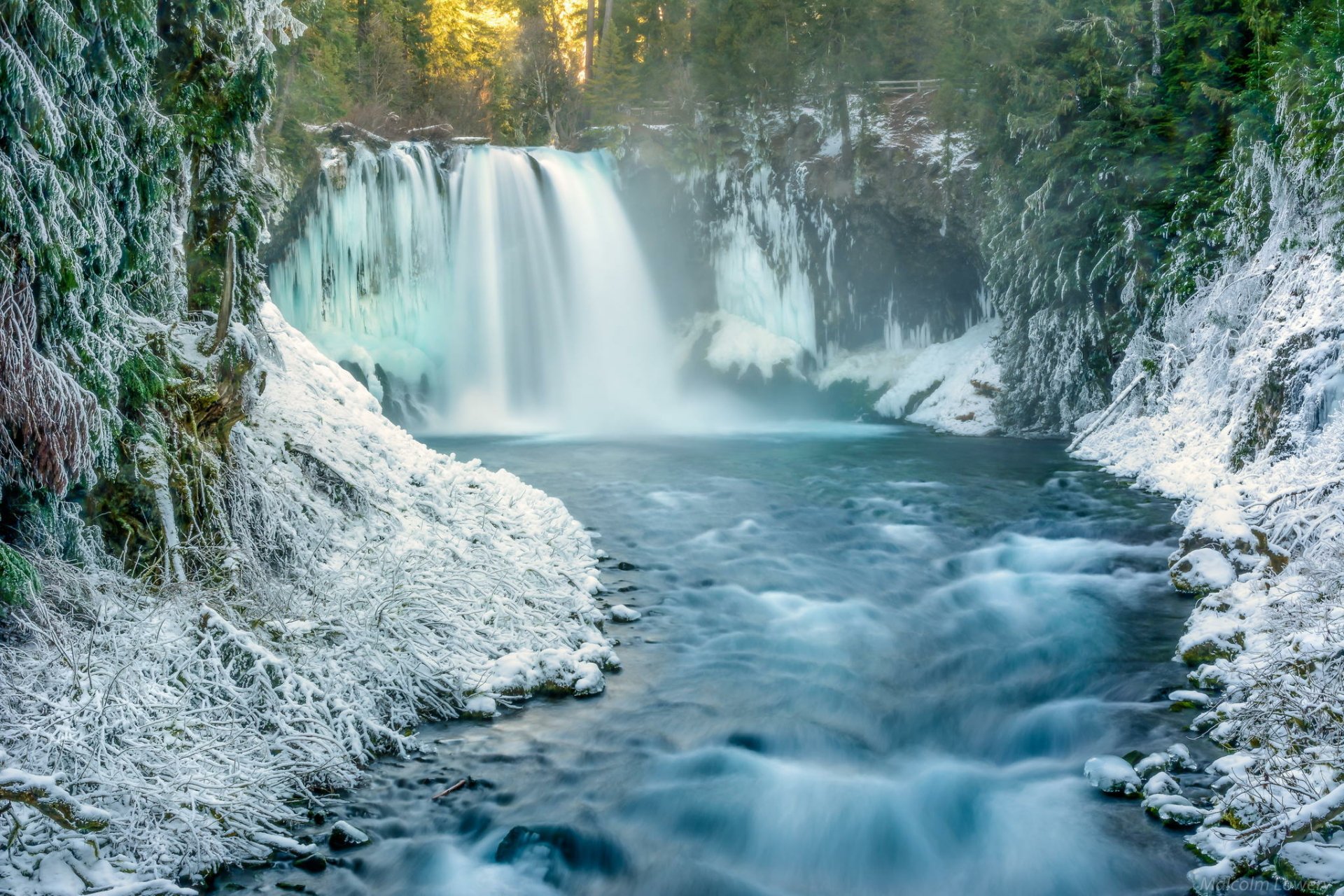 HD desktop wallpaper of a winter forest landscape featuring green trees, snow-covered ground, and a flowing waterfall surrounded by nature.