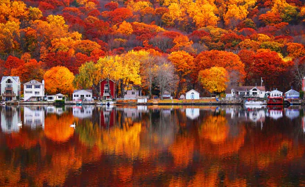 HD desktop wallpaper of a tranquil village beside a lake, surrounded by vibrant fall foliage reflecting on the water. Houses and trees create a picturesque autumn scene.