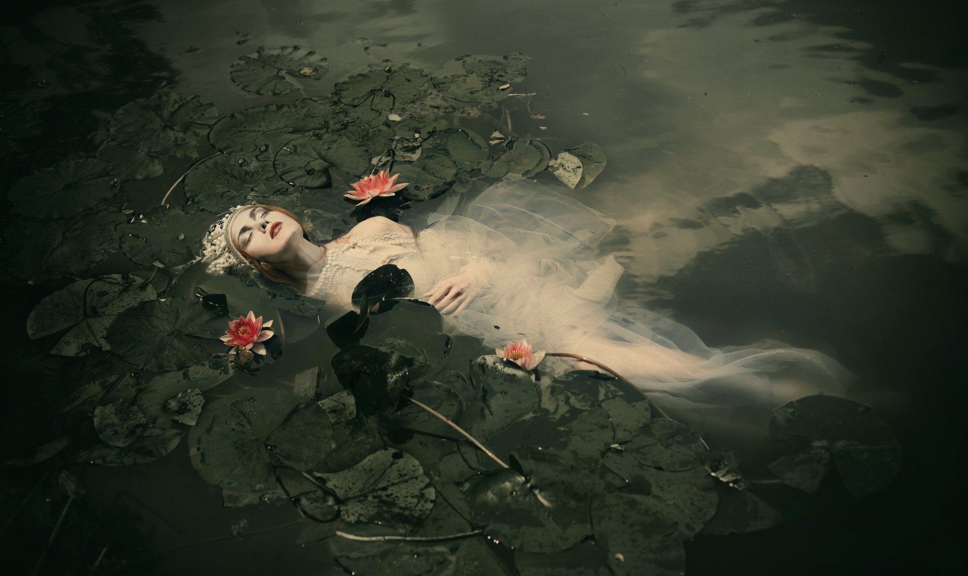 Artistic HD desktop wallpaper of a redheaded woman model floating in dark water amid lotus leaves, red lipstick and a moody, ethereal atmosphere.
