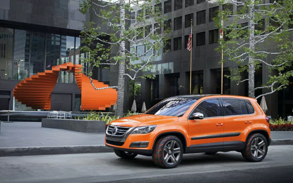 Orange Volkswagen Tiguan SUV parked on a city street with modern buildings and a matching orange spiral staircase sculpture in the background, HD desktop wallpaper.