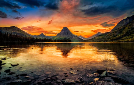 A stunning sunset over Glacier National Park, with vibrant colors reflecting on a tranquil lake surrounded by mountains, creating a breathtaking natural landscape.