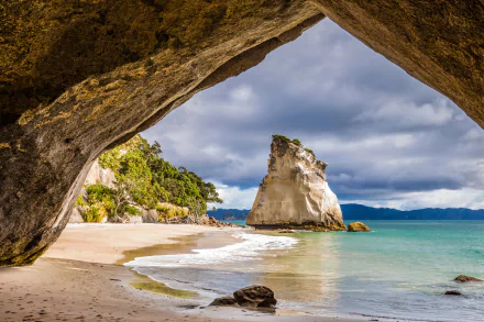 A stunning view of a coastal arch in New Zealand, framed by golden sand and lush greenery, with the ocean's gentle waves lapping at the shore under a dramatic sky.