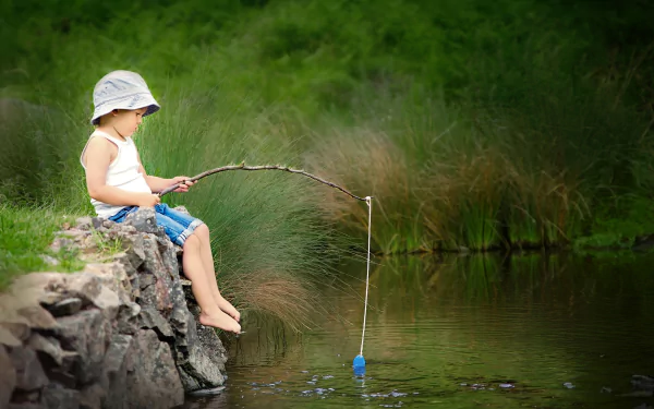 A child wearing a hat and shorts sits on a stone edge, fishing with a stick in a calm river surrounded by lush green summer foliage.