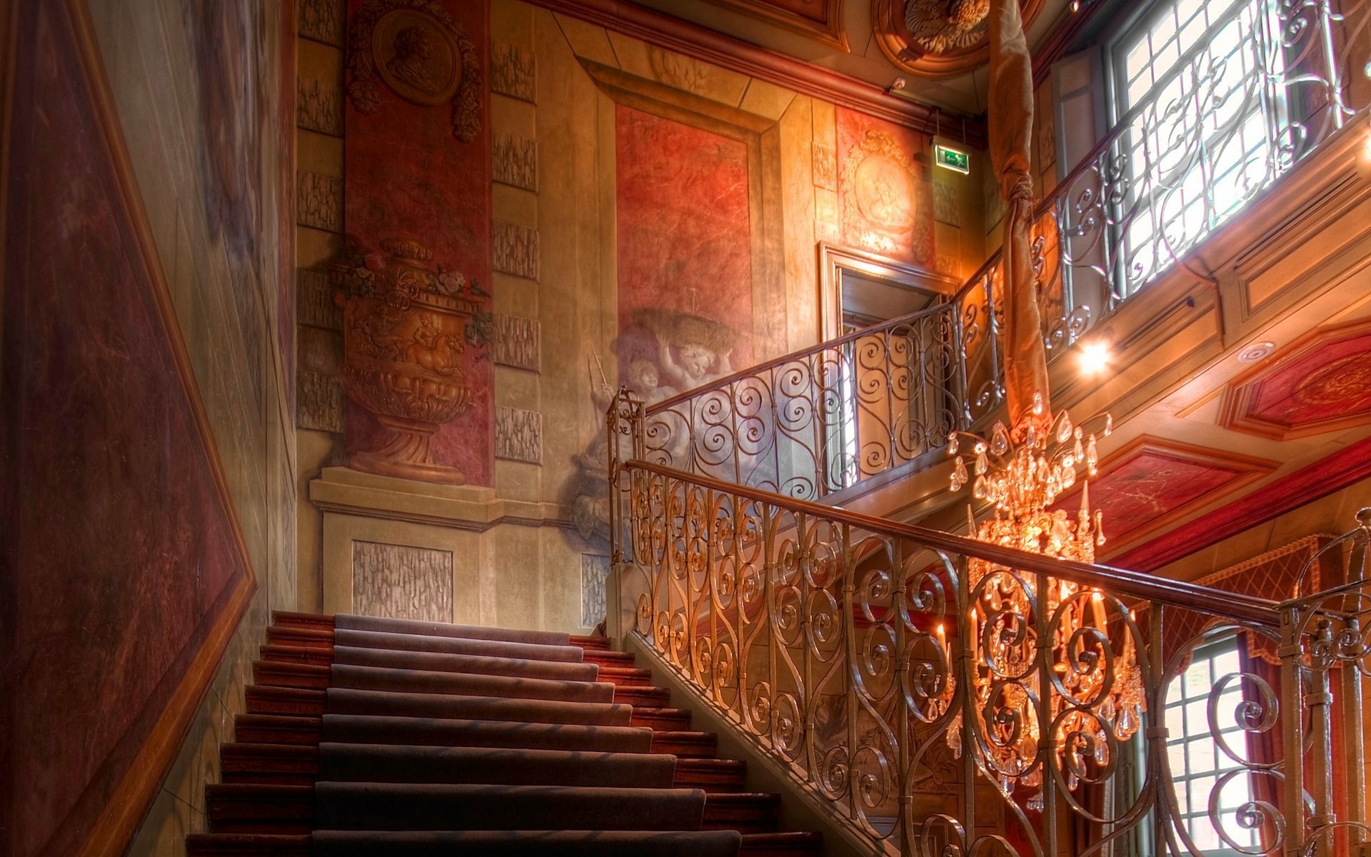 HD desktop wallpaper of an ornate colorful man-made staircase with decorative wrought-iron railing and a glowing chandelier in a richly painted interior background.