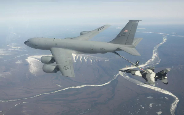 A Boeing KC-135 Stratotanker refuels a General Dynamics F-16 Fighting Falcon jet fighter in flight over a coastal landscape.